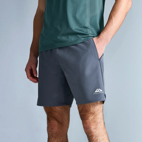 A man wearing dark gray athletic shorts with a white logo on the lower left leg, paired with a green t-shirt and gray running shoes, standing against a light gray studio background.